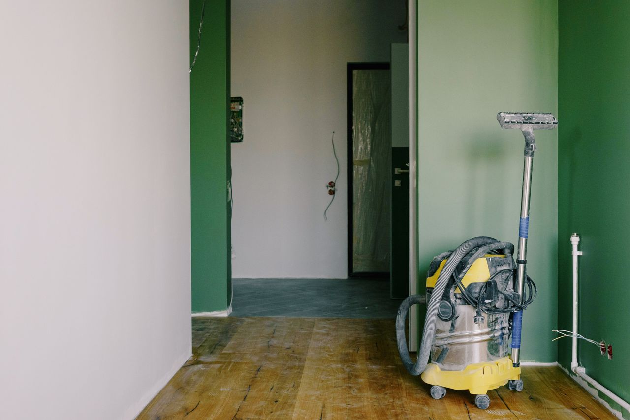 Aspirateur industriel posé sur un parquet sale dans une pièce aux murs verts et avec une porte ouverte, dans un appartement spacieux en cours de rénovation